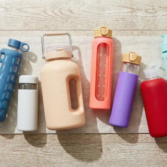 A flat lay of ten glass water bottles with colorful silicone sleeves arranged on a rustic wooden surface with natural morning light and green herbs beside them
