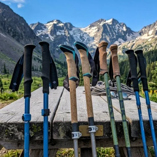 Multiple best hiking poles including Black Diamond and Leki models with foam and cork grips leaning on a wooden picnic table with a dramatic snow-capped mountain valley and wildflowers in the background